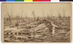 Two women are walking among the debris, another woman is sitting down. The trees have lost their leaves in the storm. Very little is recognizable among the piles of rubble except a chair and a wagon wheel.