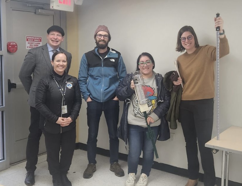 Photo shows five people standing with two of them holding a rain gauge and a snow stake.