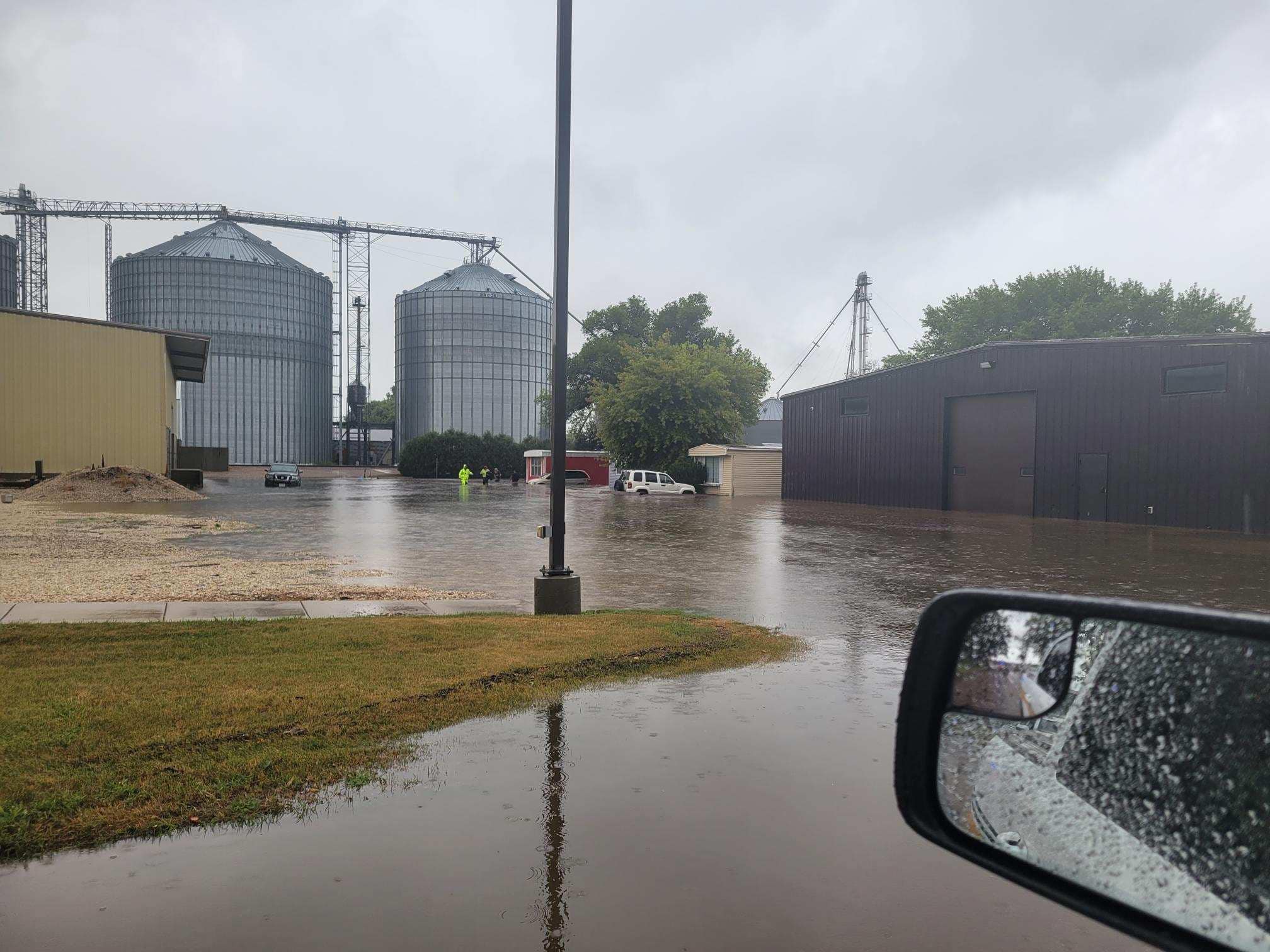 Photo taken from a vehicle in Sioux Center, Iowa showing a flooded farm. In the distance, water is up to the tops of tires on multiple vehicles parked in front of manufactured homes and other buildings. People are walking through water up to waist deep.