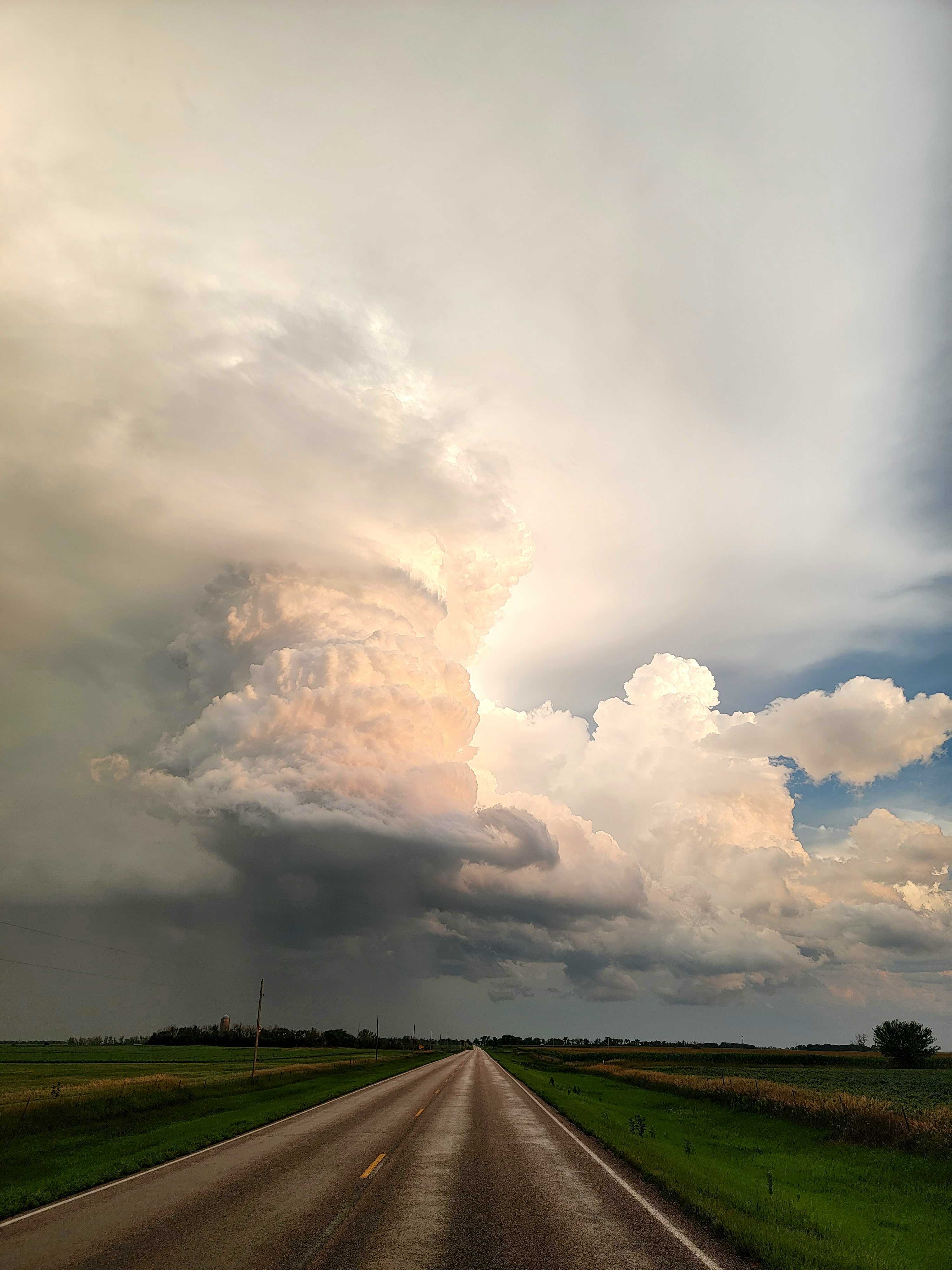 Road in the foreground stretches back through the center of the image. Above this, there is a well defined rotating column of the storm with a clear base. High clouds are above this well defined column.