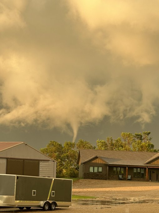 Home, garage, and trailer in the foreground. A tornado is in the center of the image, descending from a white cloud base into trees behind the house.