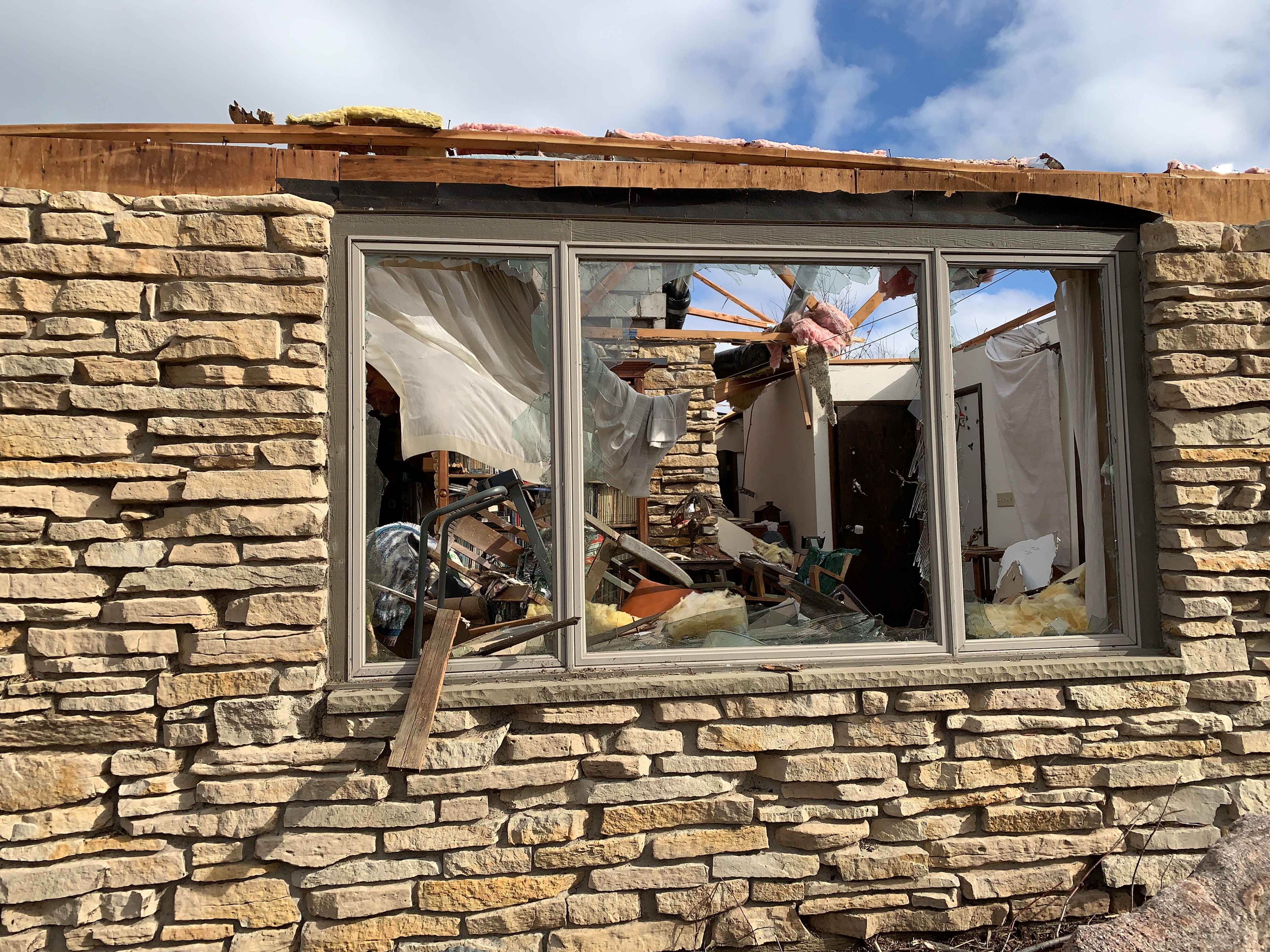 Looking through broken front window of house with roof blown off. Extensive damage in living room.